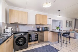 A modern kitchen with wooden cabinets, a built-in washing machine, and a bar area. - Haus 189 Dainfern- Pool  Play Area Power Backup (Sandton)