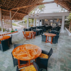 An outdoor dining area with round wooden tables and wicker chairs, surrounded by a covered patio with a thatched roof. - Hotel La Ardilla de Santa Fe (Santa Fe de Antioquia)