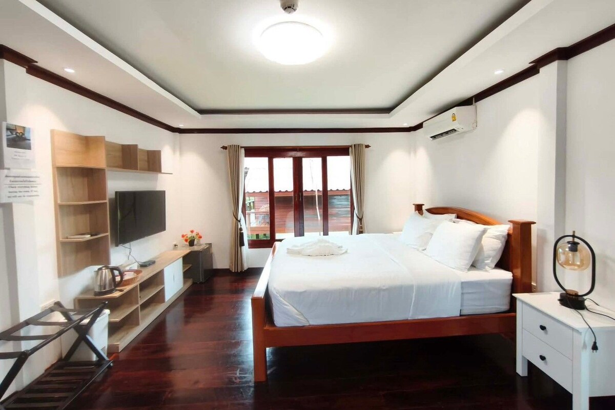 A hotel room with a bed, bedside table, TV, wooden shelving, and a view of the outside.