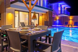 Espaço de refeições à beira da piscina, com mesa posta para uma refeição, mobiliário de exterior e piscina iluminada com iluminação ambiente.
