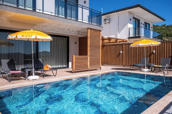 Piscina com espreguiçadeiras e guarda-sóis, cerca de madeira e um edifício com varandas.
