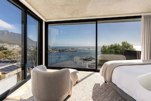 A modern hotel room with a large sliding glass door leading to a balcony with a view of the ocean and a rocky shoreline. - Ocean Villa - Iconinc Views, Perfect Location (Cape Town)