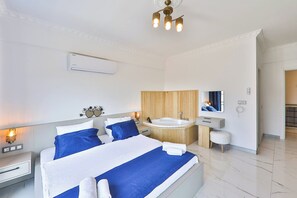 A modern bedroom with a large bed, a wooden panel wall, a built-in bathtub, and a small white stool. - villa çınar-kaş Antalya (Kaş)