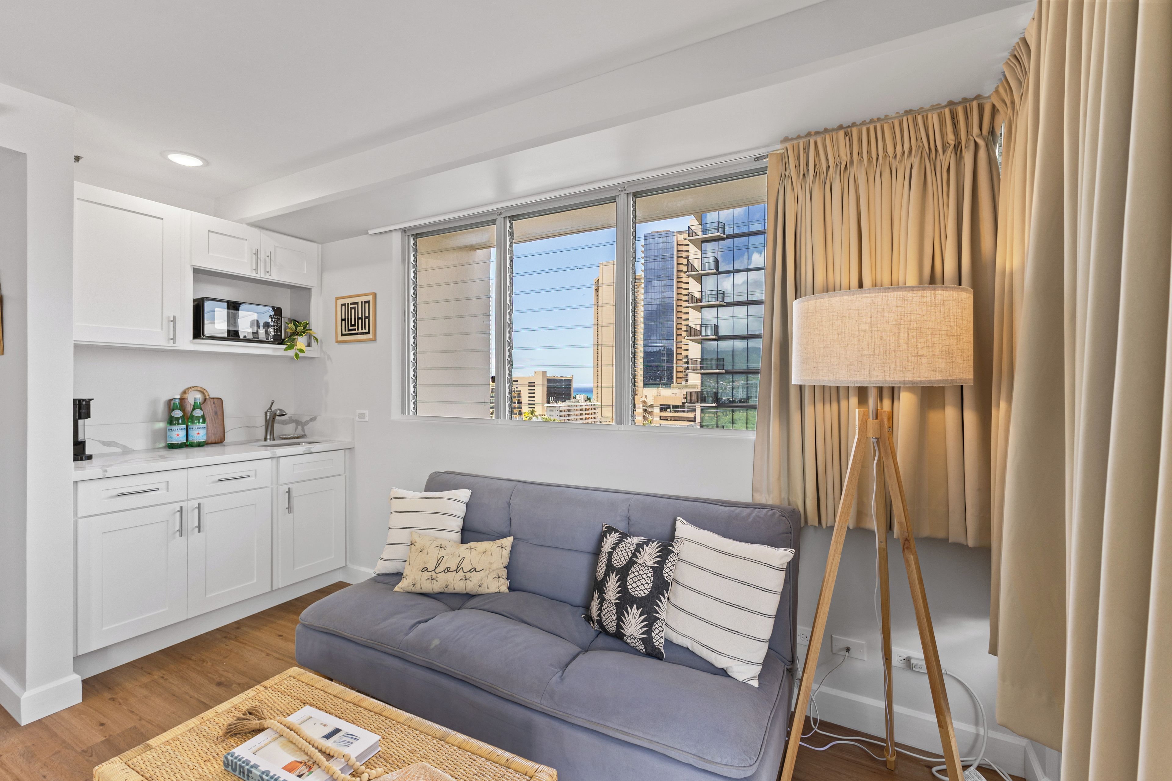 Una sala de estar moderna con un sofá gris, armarios blancos, una zona de cocina y vistas a edificios de la ciudad.