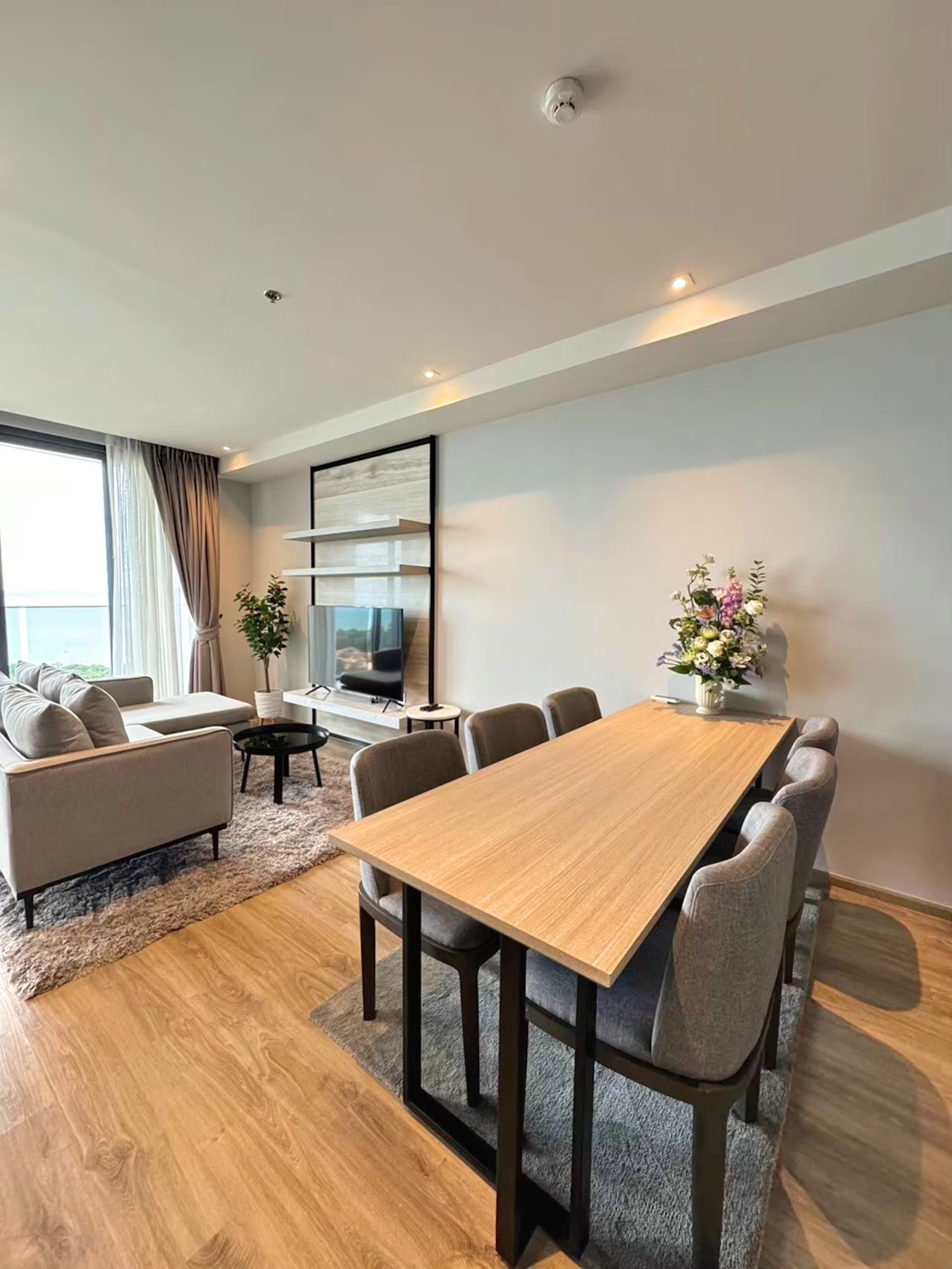Un salon moderne avec une table à manger en bois, des canapés gris, un téléviseur à écran plat et une grande fenêtre agrémentée de rideaux.