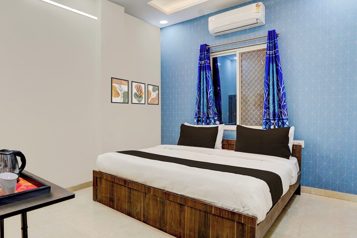 Una habitación de hotel con una cama, una mesita de noche con hervidor y una ventana con cortinas azules.