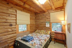 Ein Schlafzimmer in einer Blockhütte mit einem Bett, zwei Fenstern mit Jalousien, einem Nachttisch und einer Lampe.