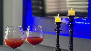 Zwei Gläser mit roter Flüssigkeit auf einer spiegelnden Oberfläche, verziert mit Tannenzapfen und getrockneten Blumen, ein Kerzenhalter mit Kerze und ein Fernseher im Hintergrund.