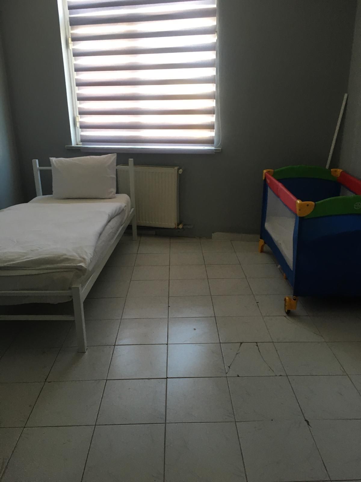 Une pièce avec un lit, un radiateur, une fenêtre avec des stores et une boîte à jouets colorée.