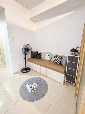 Un salon moderne avec un canapé beige, des coussins à motifs, un ventilateur sur pied, un petit tapis rond et une plante posée sur une table d’appoint.