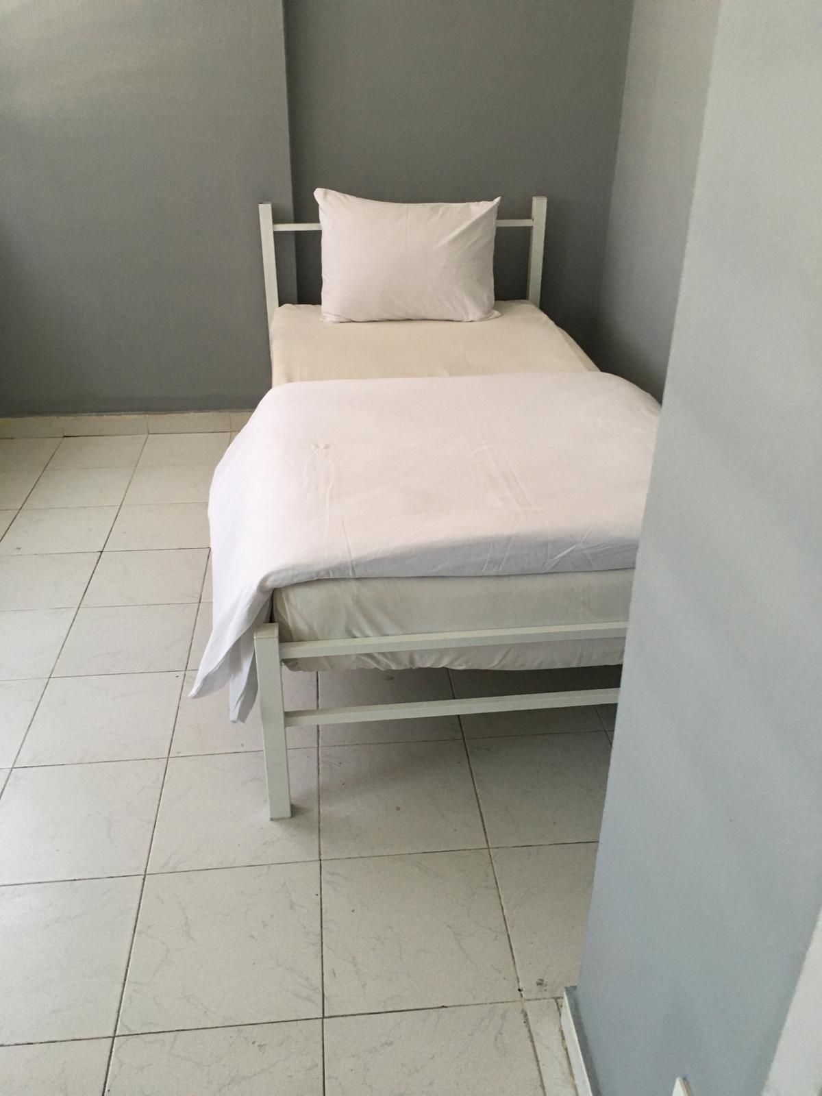 A single bed with a white mattress and pillow in a room with grey walls and tiled flooring.