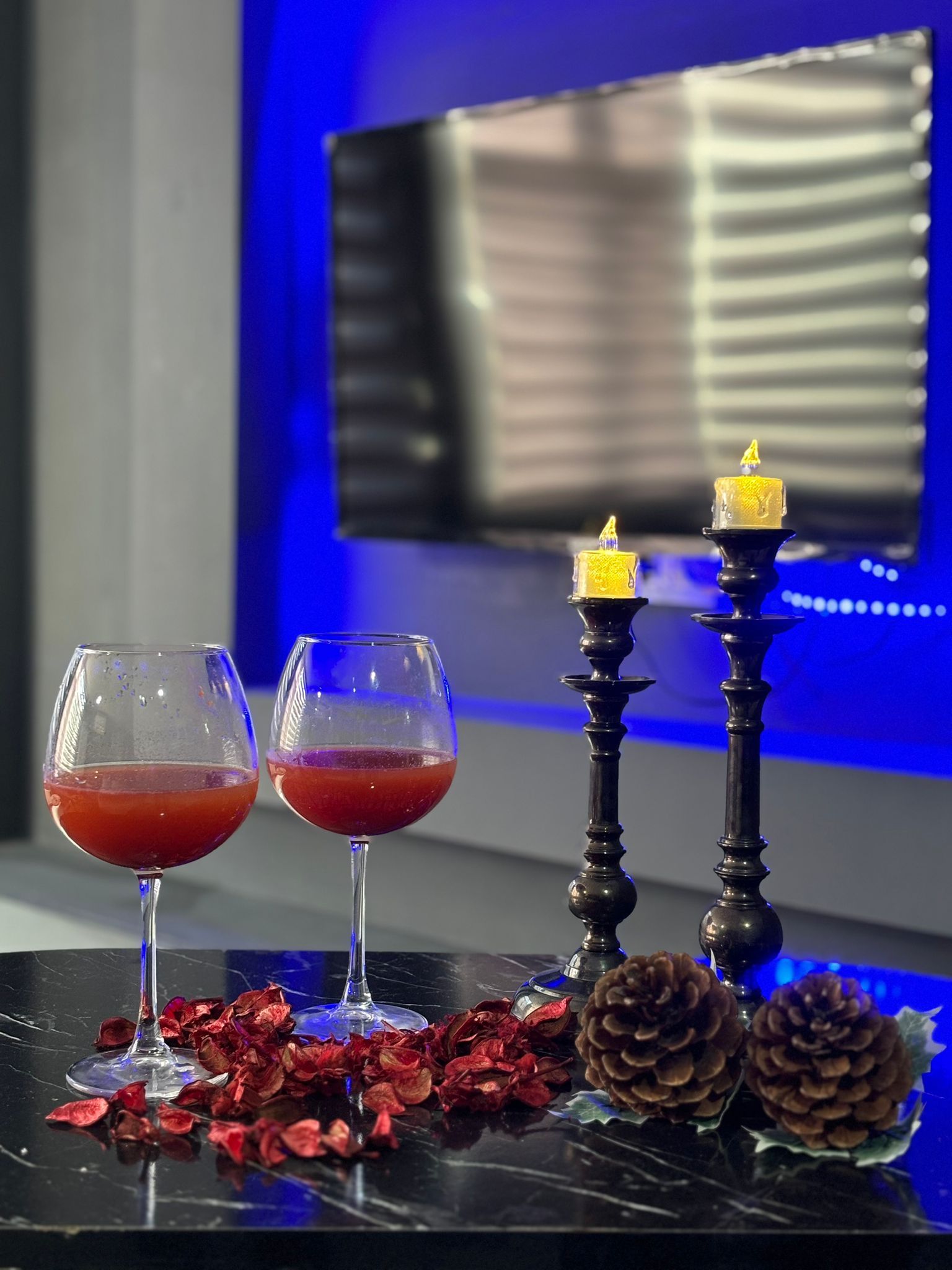 Deux verres contenant un liquide rouge sont posés sur une surface réfléchissante, accompagnés de pommes de pin décoratives et de fleurs séchées. On aperçoit également un chandelier avec une bougie et, au fond, un téléviseur.