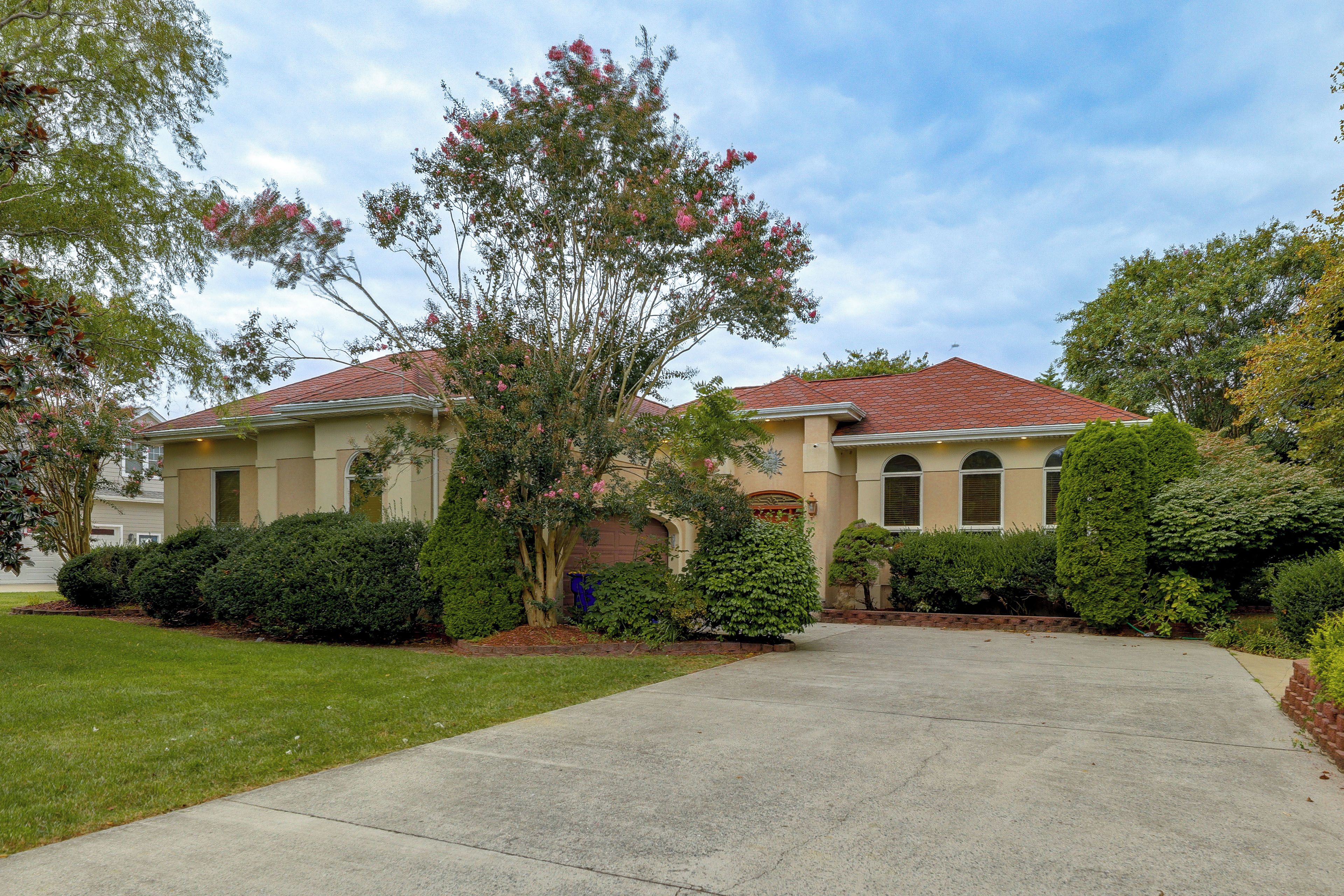 A two-story house with a red-tiled roof, a front yard with a concrete driveway, and mature landscaping including a large tree and well-maintained bushes.