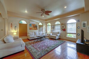 A spacious living room with a large window, a ceiling fan, and a flat-screen TV.