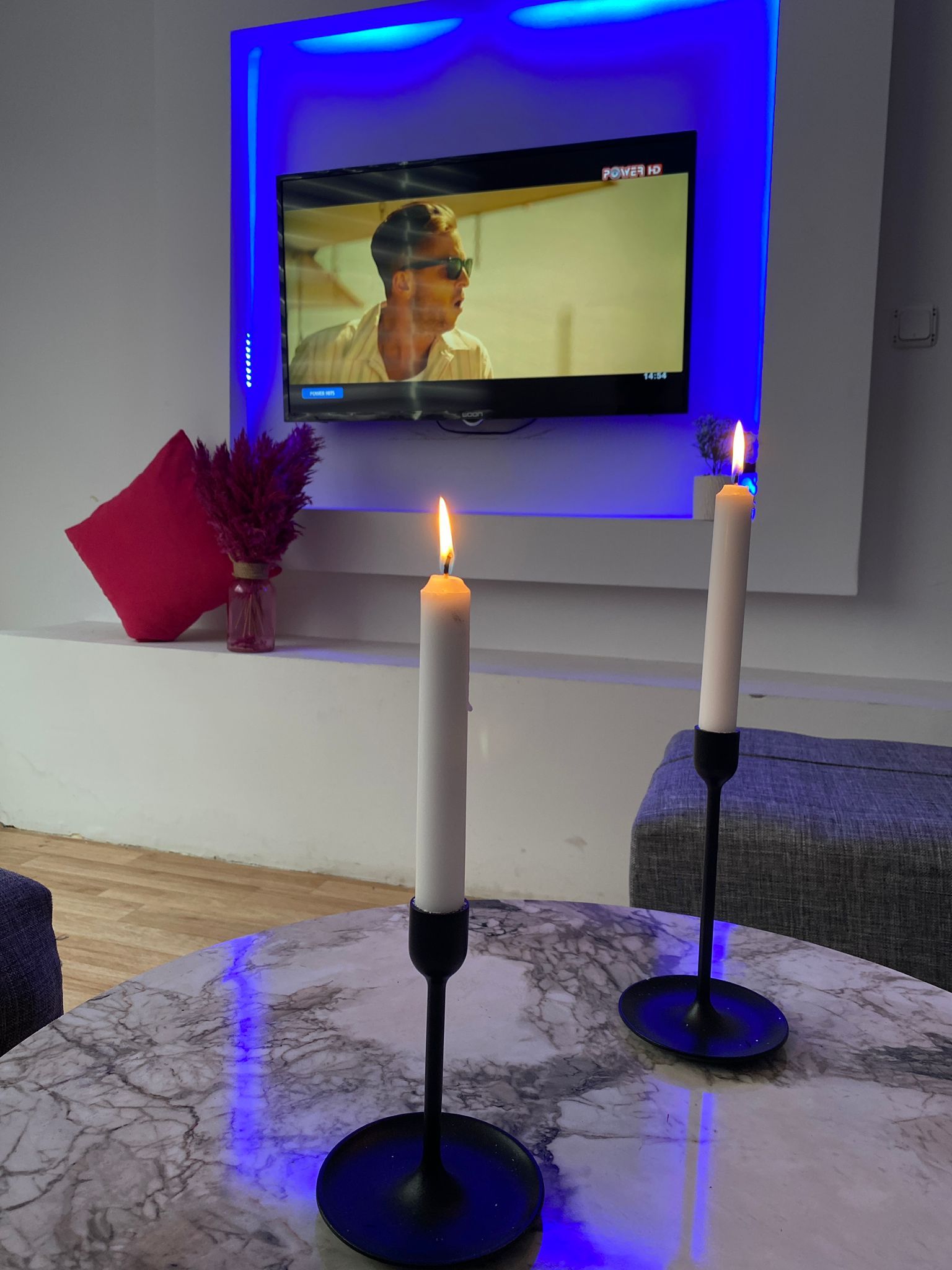 Un téléviseur à écran plat fixé au mur, avec un éclairage arrière bleu, deux bougies blanches posées sur une table en marbre et un coussin rouge sur un banc blanc.