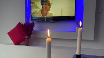 Un téléviseur à écran plat fixé au mur, avec un éclairage arrière bleu, deux bougies blanches posées sur une table en marbre et un coussin rouge sur un banc blanc.