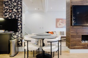 A modern living room with a round table and white chairs, a TV mounted on the wall, and a fireplace with a stone surround. - Luxury 3BDR Chelsea Townhouse Sleeps 6 (London)