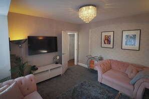 A living room with a flat-screen TV, a pink sofa, a chandelier, and two framed artworks on the wall. - Beautiful House 5 mins walk into Ulverston (Westmorland and Furness)