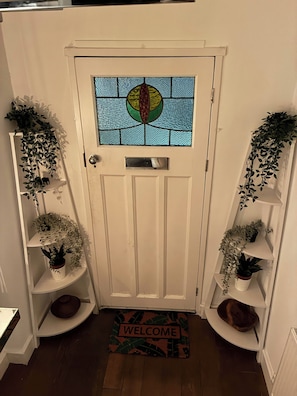 A white door with a stained glass window, flanked by white shelving units with potted plants. A welcome mat is placed at the entrance. - Beautiful House 5 mins walk into Ulverston (Westmorland and Furness)