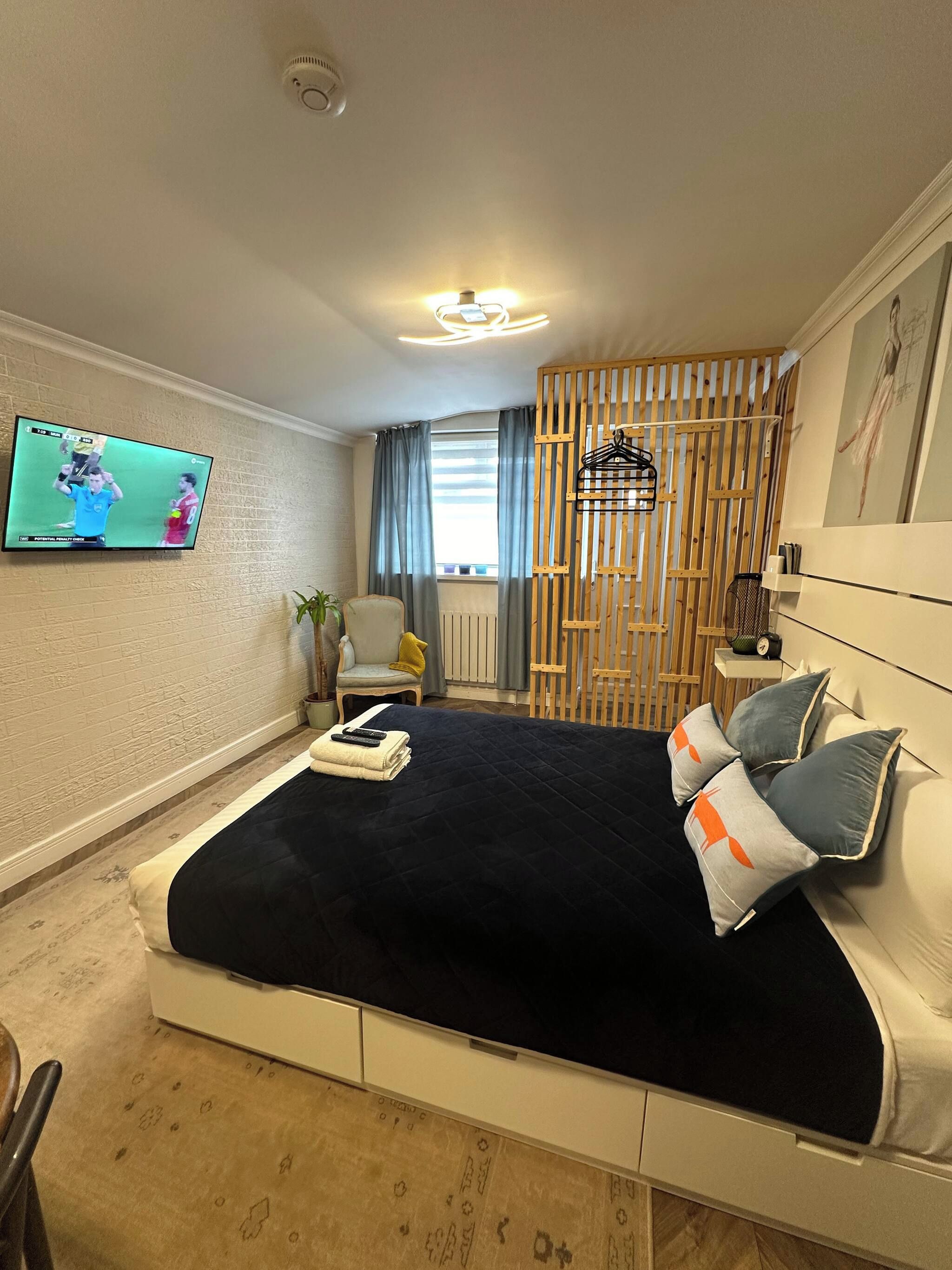 Une chambre moderne comprenant un lit, un téléviseur fixé au mur, une chaise, une petite table et un élément de séparation décoratif en bois.
