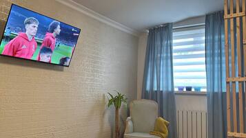Quarto de hotel com cama, televisão na parede, cadeira, planta e janela com cortinas.