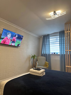 Ett hotellrum med en säng, en tv som är monterad på väggen, en stol, en växt och ett fönster med gardiner.