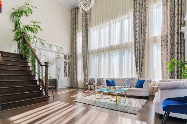 A spacious living room with a staircase, large windows with sheer curtains, a sofa set, and a coffee table.