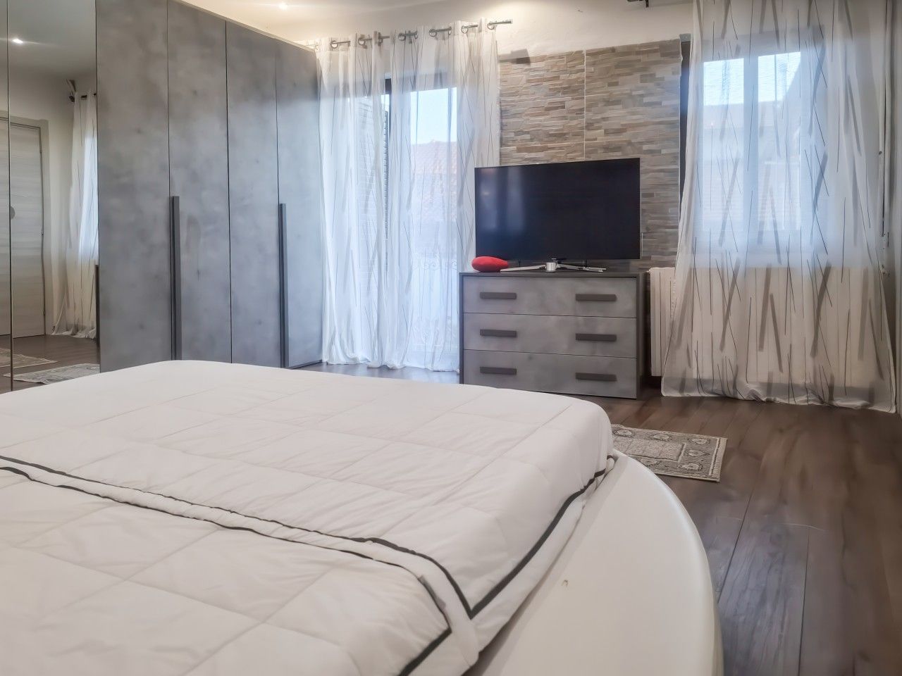 Une chambre à coucher avec un grand lit, un téléviseur à écran plat, une commode et une fenêtre ornée de rideaux légers.