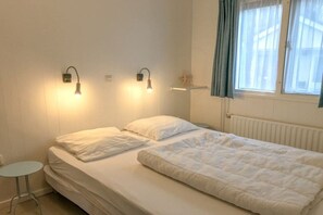 A bedroom with a bed, two wall-mounted lights, a small table, and a window with curtains. - Type A "Zee, Bos en Duin" Zoutelande (Zoutelande)