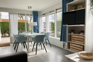 A dining area with a table and chairs, a television on a shelf, and a view of the outdoors. - Type A "Zee, Bos en Duin" Zoutelande (Zoutelande)