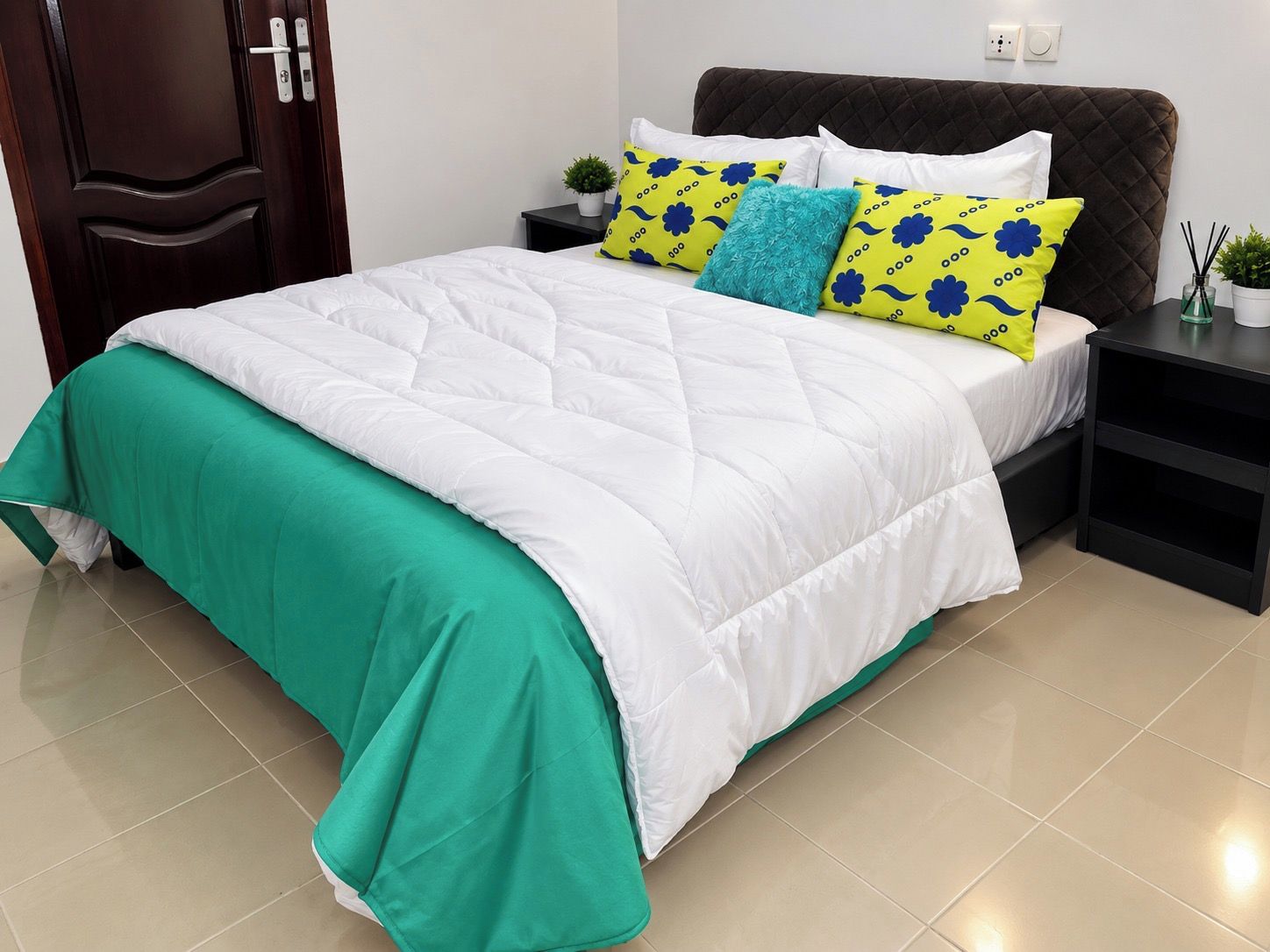 A neatly made bed with a green and white duvet, a dark headboard, and a bedside table with a small plant and a lamp.