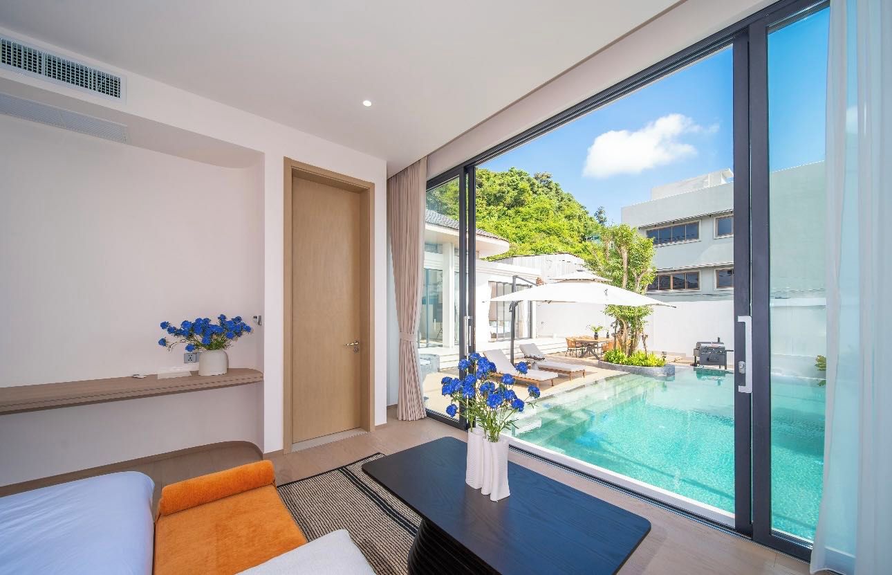 Una habitación de hotel moderna con un ventanal grande, una cama, una mesita con flores y vistas a una piscina y edificios.