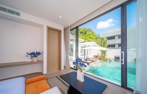 A modern hotel room with a large window, a bed, a small table with flowers, and a view of a pool and buildings. - Kata beach 4bedroom luxury pool villa (Karon)