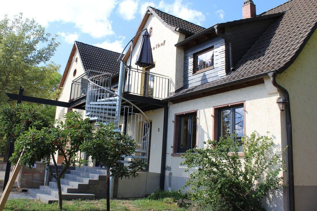 Ein zweistöckiges Haus mit einer Wendeltreppe, die zu einem Balkon führt, umgeben von viel Grün.