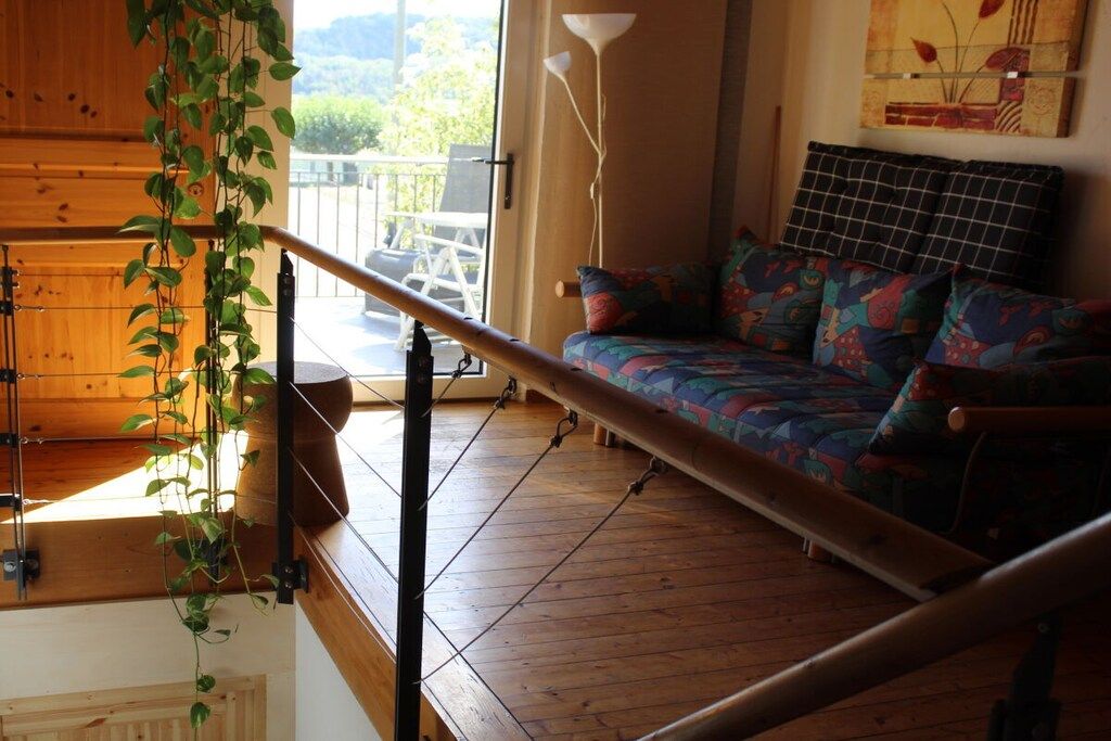 Ein Zimmer mit Holzboden, einem farbenfrohen Sofa, einer hängenden Pflanze und Blick durch ein Fenster.