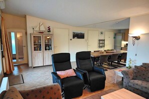 A living room with a sofa, two reclining chairs, a dining table with chairs, a cabinet with glass doors, and a kitchen area in the background. - Holiday home with 3 bedrooms (Westkapelle)