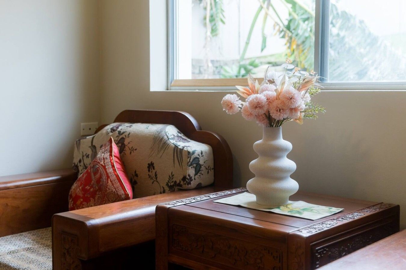 Une table en bois sur laquelle se trouve un vase blanc contenant des fleurs roses, un coussin à motifs et un sac rouge.