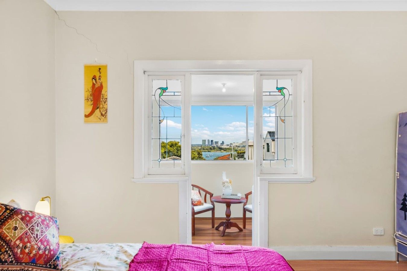 A room with a bed, a table with chairs, a window with a view of a cityscape, and a painting on the wall.