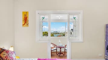 A room with a bed, a table with chairs, a window with a view of a cityscape, and a painting on the wall.