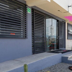 A modern building facade with horizontal slats, a green emergency exit sign, and a visible entrance with a glass door. - Estancias Vive MX el Marques (El Marques)