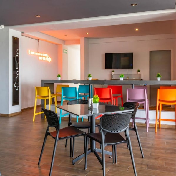 A modern dining area with a central table and colorful chairs, a flat-screen TV mounted on the wall, and a neon sign on the wall. - Estancias Vive MX el Marques (El Marques)