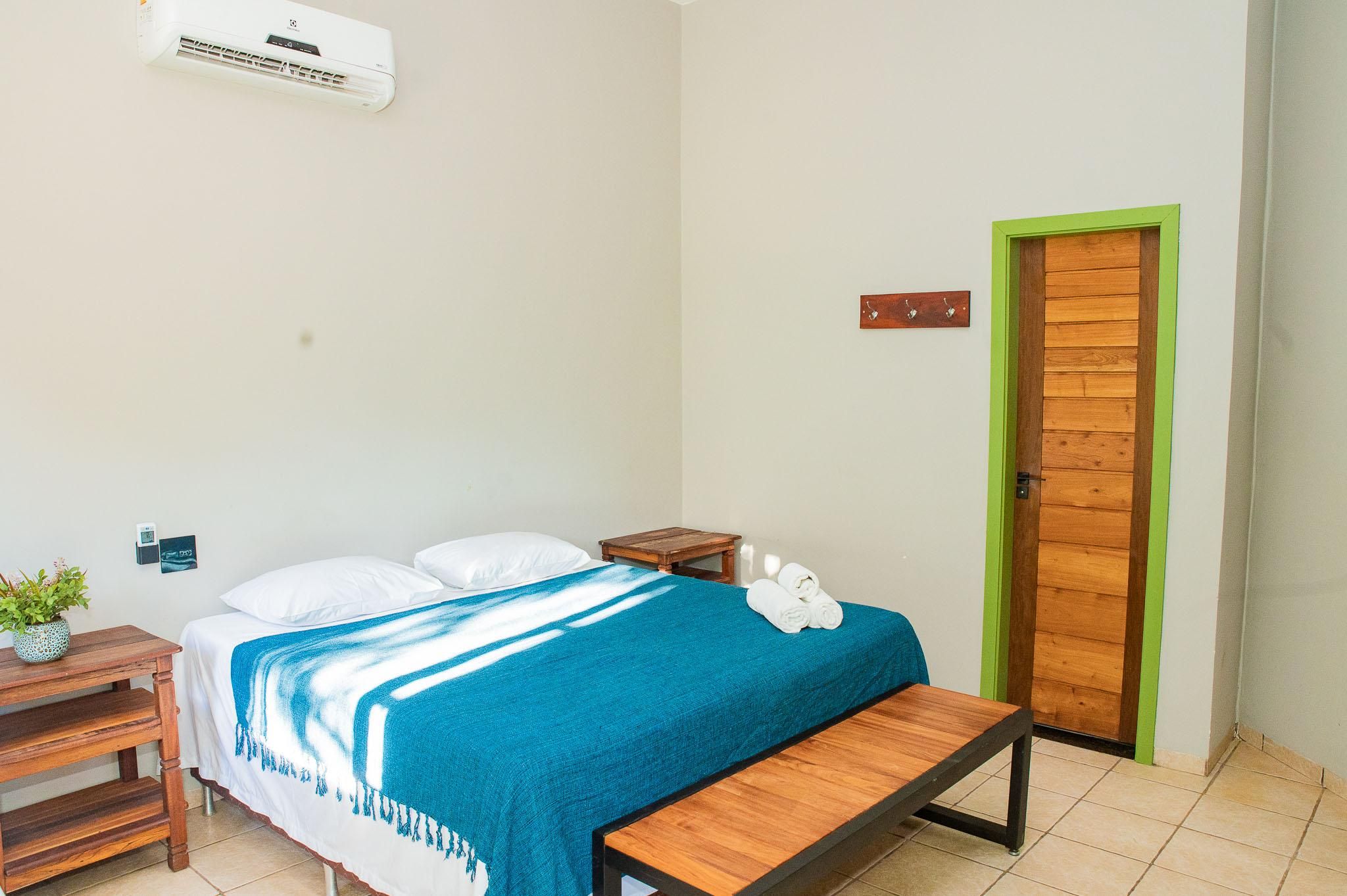 A hotel room with a bed, a bench, a small table, a wall-mounted air conditioner, and a door with a green frame.