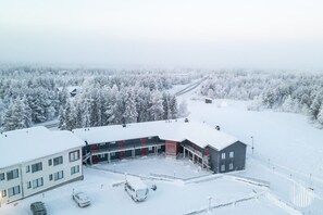 Ein mehrstöckiges Gebäude mit Parkplatz und verschneiter Landschaft, Bäume im Hintergrund.