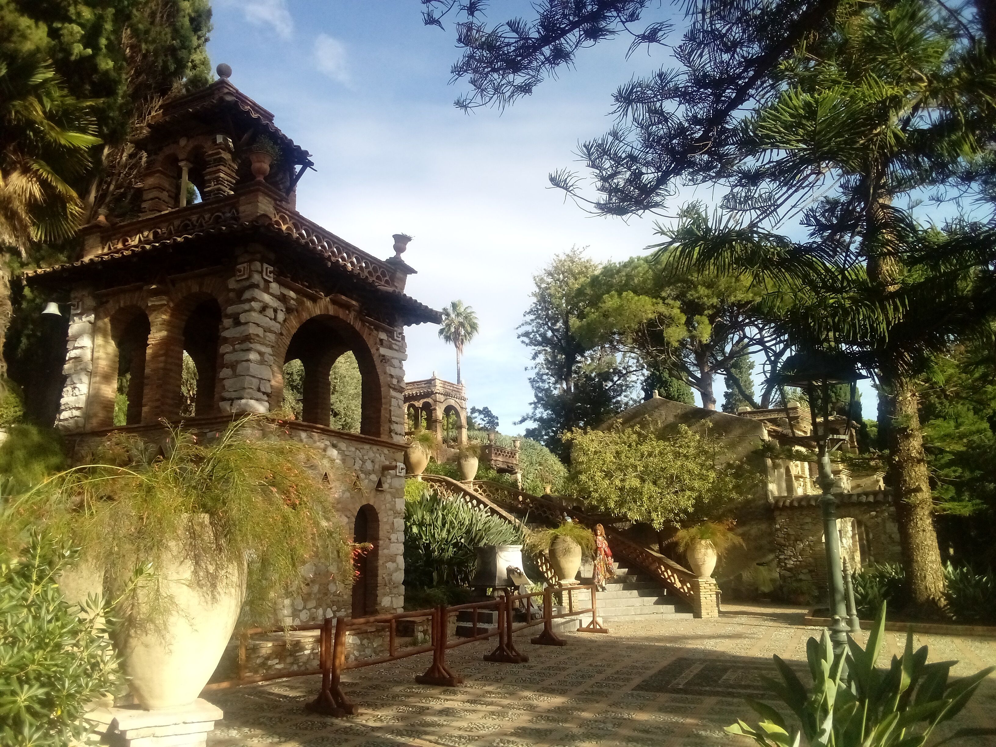 A stone structure with arches and a tiled roof, surrounded by potted plants and trees.