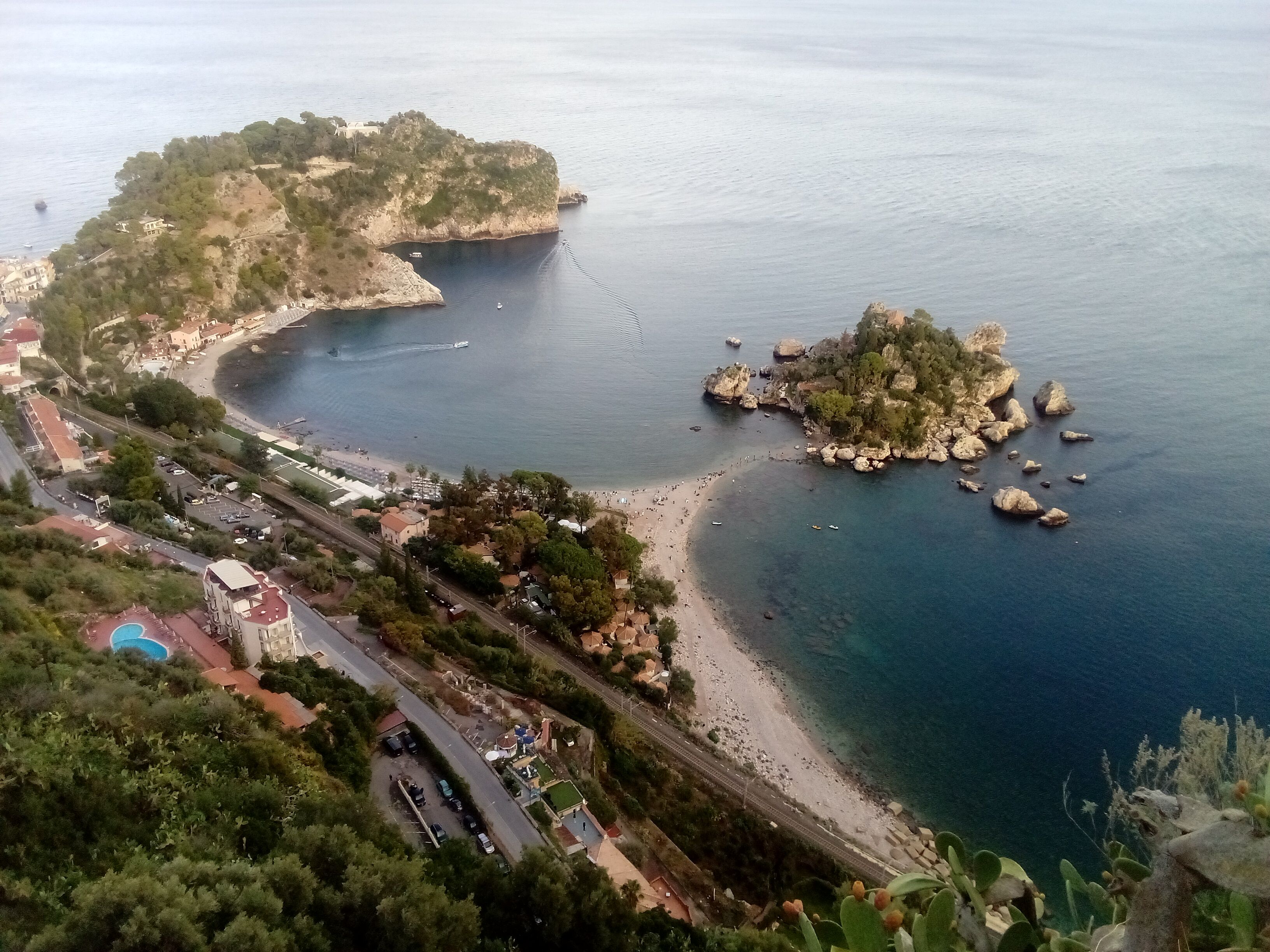 A coastal landscape with a beach, rocky outcrops, and a road leading to a building with a pool.