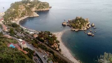 A coastal landscape with a beach, rocky outcrops, and a road leading to a building with a pool.