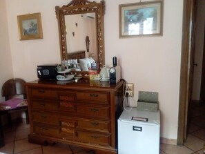 Uma cômoda de madeira com gavetas, um pequeno frigorífico, uma máquina de café e uma chaleira em cima. Há quadros nas paredes e um espelho acima da cômoda.