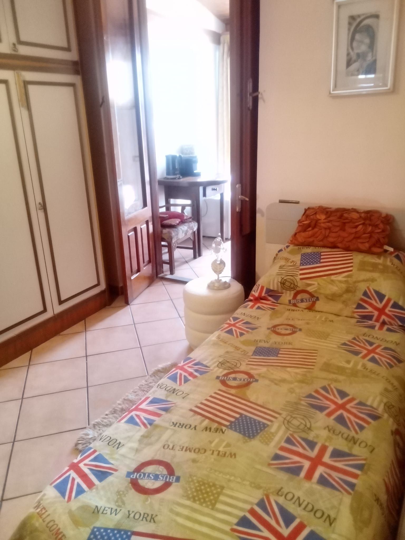 A bedroom with a bed featuring a quilt with flags, a chair, a small table, and a door leading to another room.