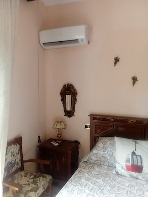 Quarto de hotel com cama, mesa de cabeceira com luminária, cadeira, ar-condicionado fixado na parede e espelho decorativo.