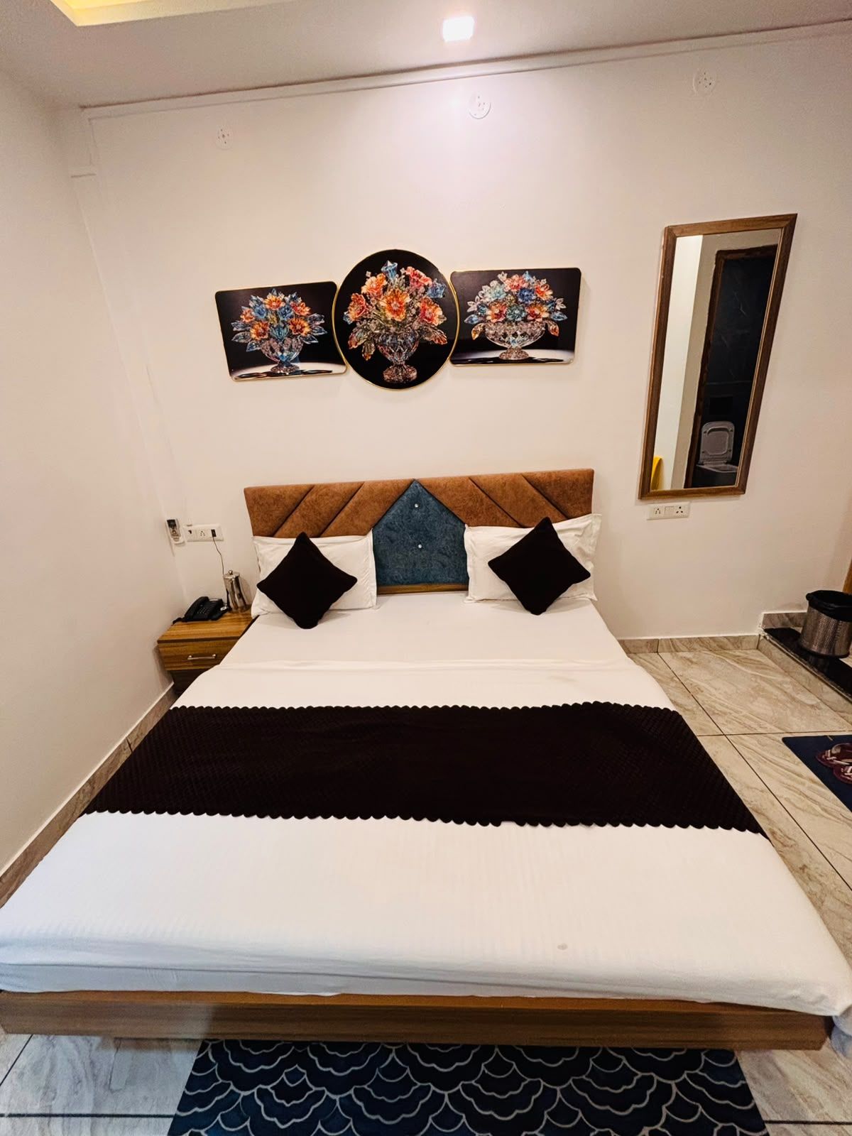 Una cama bien tendida con cabecera de madera, una mesita de noche con lámpara y un espejo montado en la pared.